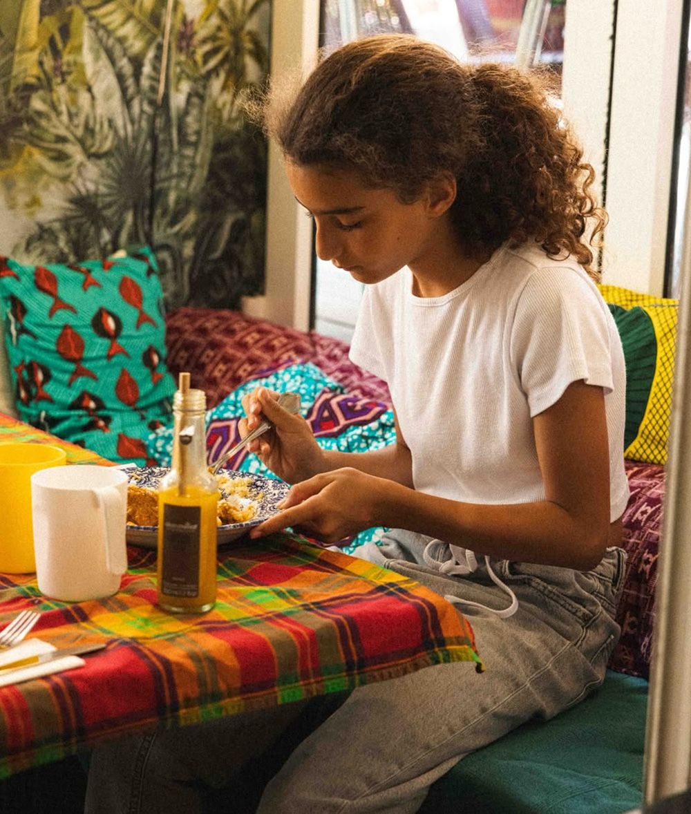 Une jeune personne aux cheveux bouclés prend un repas à une table colorée et confortable avec des coussins à motifs. Limbus Studio