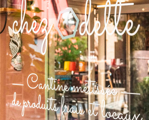 Porte en verre avec écriture blanche indiquant "chez Odette, Cantine métissée de produits frais et locaux", reflétant l'intérieur d'un café. Pour en savoir plus sur chez odette, consultez le site chezodette.com. Limbus Studio