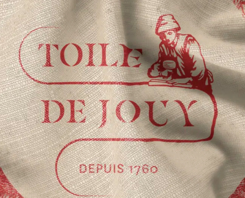 Tissu beige avec impression rouge indiquant "Toile de Jouy" et "Depuis 1760", avec un personnage dessinant au-dessus du texte. Parfait pour les amoureux de la tradition textile française et les fans du Musée de la Toile de Jouy. Limbus Studio