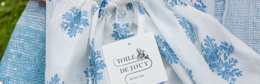 Un tissu blanc avec un imprimé floral bleu et une étiquette "Toile de Jouy" repose élégamment sur une surface texturée bleue, mettant en valeur le style classique de la Toile de Jouy. Limbus Studio