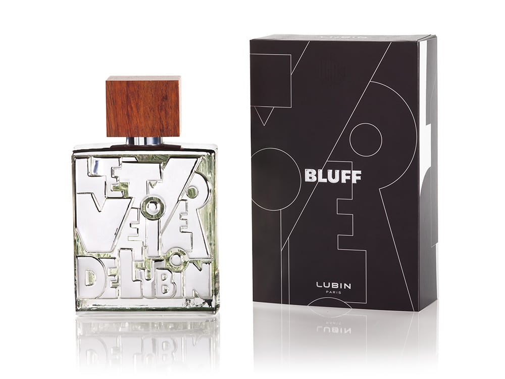 Un flacon de parfum avec un bouchon en bois et un design de texte complexe se trouve à côté d'une élégante boîte noire étiquetée « BLUFF » et « LUBIN PARIS », présentant une création magistrale d'élégance et d'art.