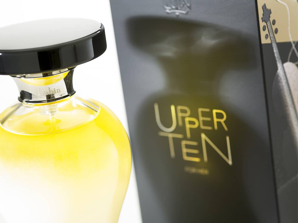 Un flacon de parfum avec un bouchon noir se trouve à côté d'une boîte étiquetée « Upper Ten » dans un élégant design doré et noir, semblable à la sophistication élégante trouvée sur un site WordPress bien conçu.