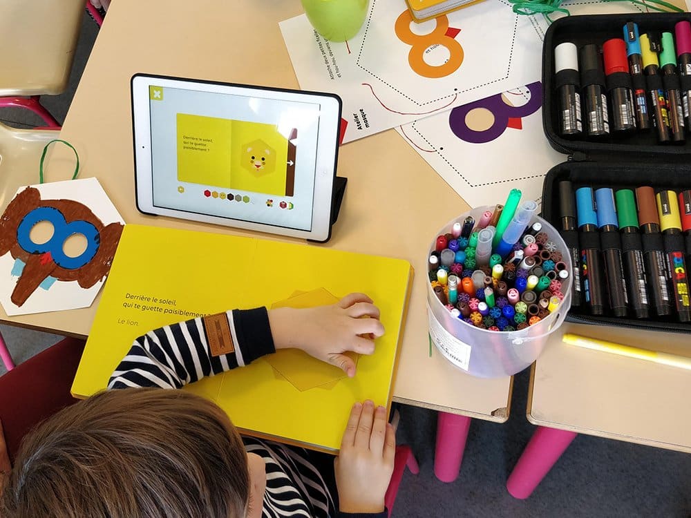 Dans un coin studio cosy, un enfant s'adonne à la création, mélangeant des merveilles numériques sur un iPad avec du papier vibrant et des marqueurs éparpillés sur la table. Limbus Studio