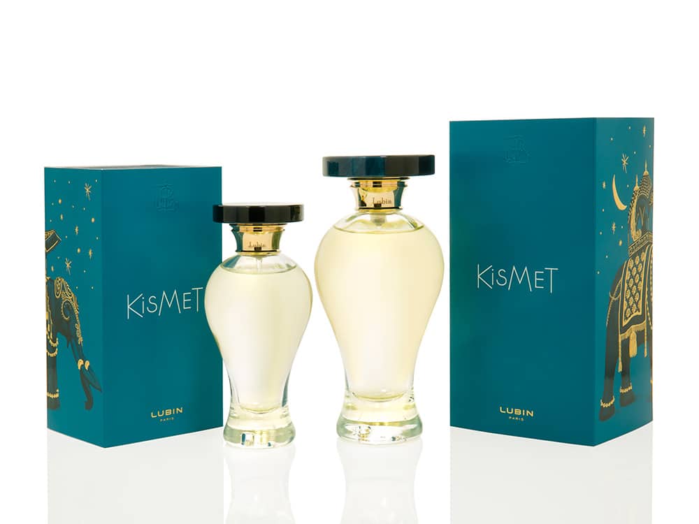 Deux élégants flacons de parfum Kismet aux bouchons noirs côtoient des boîtes bleu sarcelle ornées de motifs dorés. L'emballage raffiné est une véritable création, mêlant art et sophistication dans chaque détail.